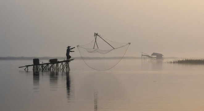Traditional fishing method with cast net on wooden pier at misty sunrise. Silhouette of fisherman catching fish on calm lake. Rural landscape in morning fog for postcard