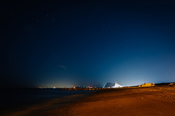 A peaceful night view of a beach with a starry sky and the illuminated Rock of Gibraltar in the background, creating a serene atmosphere..