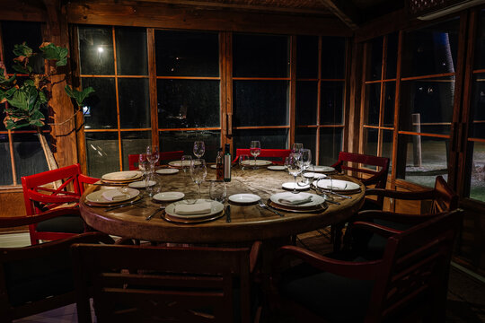 A rustic dining setup with a round wooden table, elegant tableware, wine glasses, and red chairs in a cozy, warmly lit indoor space.