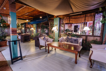 A stylish and cozy indoor lounge with wooden furniture, white armchairs, warm lighting, and lush green plants, creating a relaxing ambiance.
