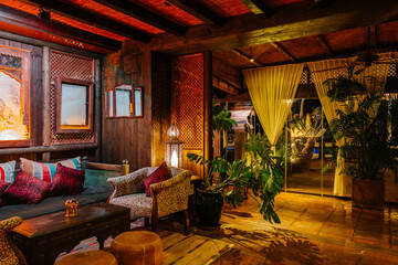 A cozy, warmly lit lounge with exotic decor, wooden furniture, patterned cushions, lush plants, and...