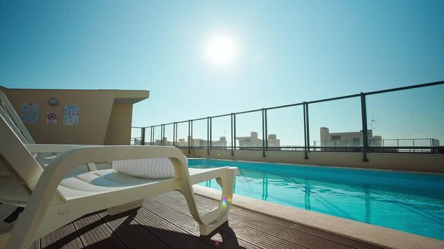 White sun loungers with rolled towels beside a rooftop pool under a bright sun, wooden deck and glass railing overlooking the city skyline. Luxury relaxation atmosphere