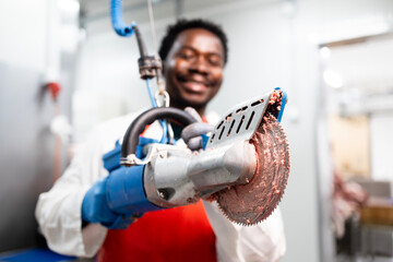 Experienced food factory worker butcher using circular electric saw to cut fresh meat in...