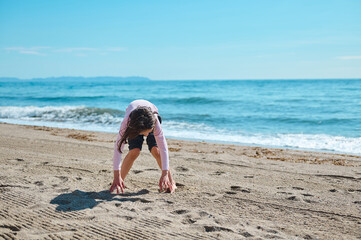 A young girl crouches on the sandy beach near the ocean under the sunshine, enjoying a summer day outdoors.