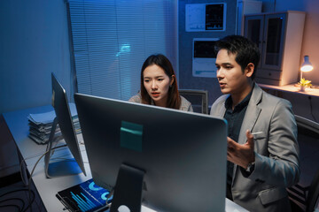 Professional asian colleagues reviewing financial charts on computer screen, working late in dimly lit corporate office with monitor's soft illumination