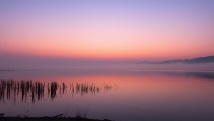 Obraz premium A serene lake at sunset, featuring a tranquil reflection of colorful skies and mist, framed by gentle reeds along the water's edge. 