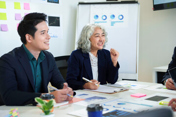 Professional colleagues collaborating, reviewing presentation materials at white desk, surrounded by documents, sticky notes, whiteboard displaying data charts