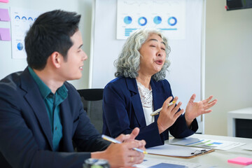 Senior businesswoman gesturing and explaining strategy to young businessman during a meeting in a conference room, with charts and graphs visible in background