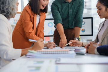 Asian business team collaborating, reviewing marketing strategy, pointing at charts and graphs during conference meeting in contemporary corporate workspace