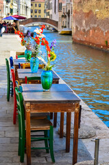 I tavolini di un ristorante sulla riva di un canale di Venezia abbelliti con dei vasi in vetro e dei fiori colorati