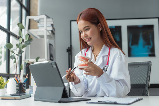 Asian female doctor wearing white coat and stethoscope is holding denture model and pointing with pen at digital tablet during video call in hospital