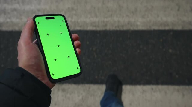 Pov view male hand holding modern smartphone keylight green screen gadget walking street at night, camera locked on cell phone chroma key display, zebra crosswalk evening illumination, copy space