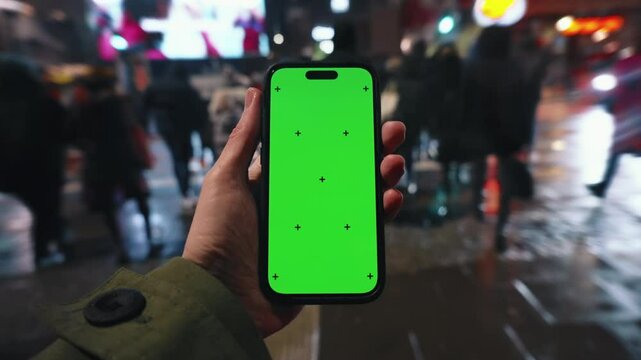 Pov look at male hand holding smartphone keylight green screen gadget in street at night, man walking crowded crosswalk, camera locked on phone chroma key display, bokeh evening city background