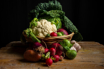 View of basket with cauliflower, cabbage, potatoes, radishes and garlic on black background table, horizontal with copy space
