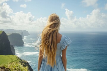 Obraz premium a blonde woman with long hair wearing a light dress, standing on a cliff and gazing at the sea