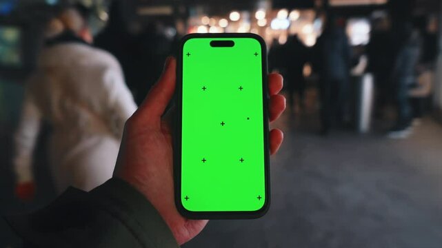 Pov look at male hand holding smartphone keylight green screen gadget in street at night, man walking crowded sidewalk, camera locked on phone chroma key display, bokeh evening city background