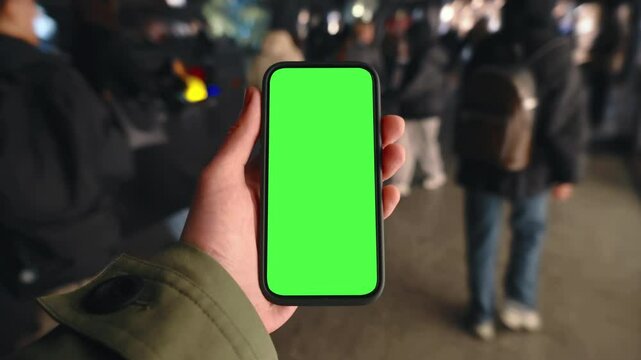 Pov look at male hand holding smartphone keylight green screen gadget in street at night, man walking crowded sidewalk, camera locked on phone chroma key display, bokeh evening city background