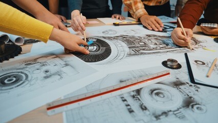 A diverse group of engineers collaborates around a table filled with detailed machinery drawings, showcasing teamwork and creativity in the engineering process. SACTR