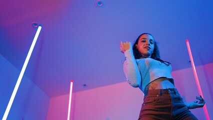 Low angle camera of hispanic dancer looking at camera and dancing to city pop music. Professional attractive hipster moving to hip hop song with purple led or neon light. Stylish cloth. Regalement.
