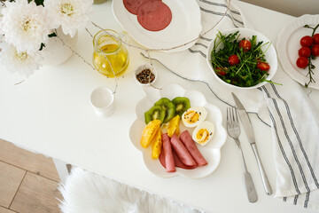 A nutritious and healthy, colorful breakfast plates features eggs, fruits, and a fresh salad wit vase with aragula, ham, olive oil elegantly arranged on a white tablecloth.