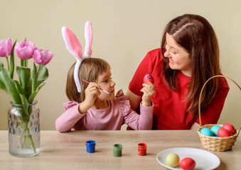 Naklejka premium Toddler girl with bunny ears and brush in one hand showing to mother red colored egg she just painted. Mom happy for her child. Basket of dyed eggs on table. Spring holiday and togetherness concept