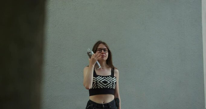 In a sunny afternoon, a woman wearing a trendy outfit stands outside a contemporary building, taking a refreshing sip of water to cool off.
