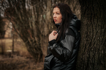 A woman in a black hooded jacket leans against a tree, gazing into the distance. The blurred forest...