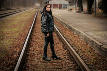 A woman in a black leather outfit and high-heeled boots stands on railway tracks, extending her...