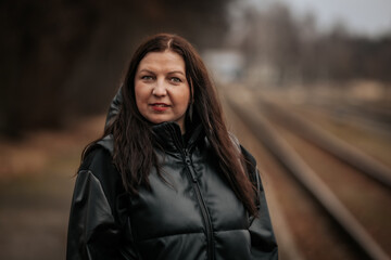 A woman in a black hooded jacket stands near train tracks, gazing at the camera with a confident...