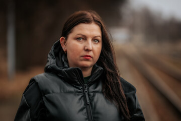 A woman in a black hooded jacket stands near train tracks, gazing at the camera with a confident...