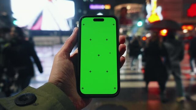 Pov looking at male hand holding smartphone keylight green screen gadget in street at night, man walking crowded crosswalk with phone chroma key display, bokeh modern city illumination in evening