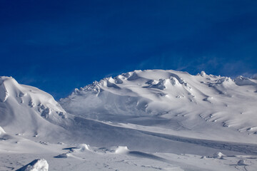 The snow-covered surface of the mountainside. The top of this mountain is exposed to strong winds that blow snowflakes away like dust against the blue sky