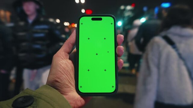 Pov looking at male hand holding smartphone keylight green screen gadget in street at night, man walking crowded crosswalk with phone chroma key display, bokeh modern city illumination in evening