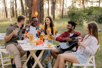 Meeting of multiracial group of friends playing guitar, singing, eating dinner and drinking wine during party in the forest