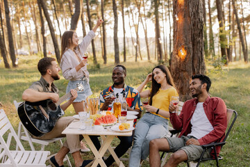 Meeting of multiracial group of friends playing guitar, singing, eating dinner and drinking wine during party in the forest