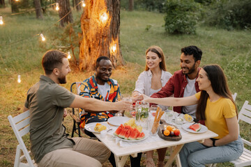 Multiracial group of happy friends sitting around table drinking wine talking at dinner party in evening summer forest