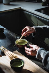 Close-up of hands using a bamboo whisk and spoon to prepare matcha tea in a beautifully lit modern kitchen setting.
