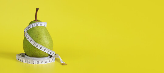 Measuring tape and green ripe pear on yellow background with space for text. Healthy eating and weight loss concept. Composition with white measuring tape and pear. Diet. Metaphor - pear figure shape