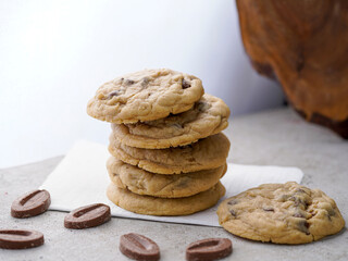 A stack of freshly baked chocolate chip cookies with a soft, chewy texture, placed on a white napkin. Surrounding the cookies are chocolate pieces, adding a delicious and tempting touch.