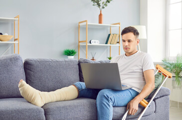Handicapped young man working on a laptop at home sitting on sofa with crutches after accident. Male injured person with a broken leg using computer on rehabilitation indoors. Rehab concept.