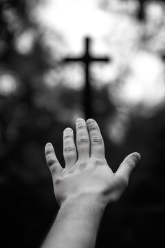 Black and white artistic shot of hand reaching toward the sky with faint cross in the blurred background. Symbolizing faith and hope. Good Friday concept.