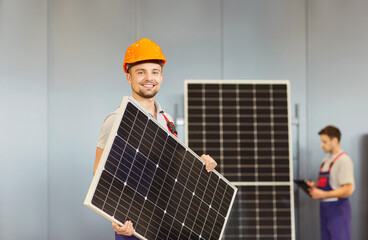 Smiling worker wearing hard hat holding a solar panel in his hands while his colleague in work...