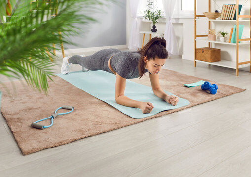 Slim athletic girl having fitness workout at home, doing plank exercise in the living room. Woman in sportswear is standing in plank position on yoga mat. Fitness, wellness and home training concept - Powered by Adobe