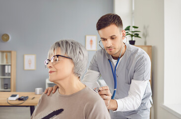 Obraz premium Health of older people. Young male doctor listens to elderly woman's heartbeat during her clinic visit. Healthcare professional using stethoscope on elderly female patient during medical examination.
