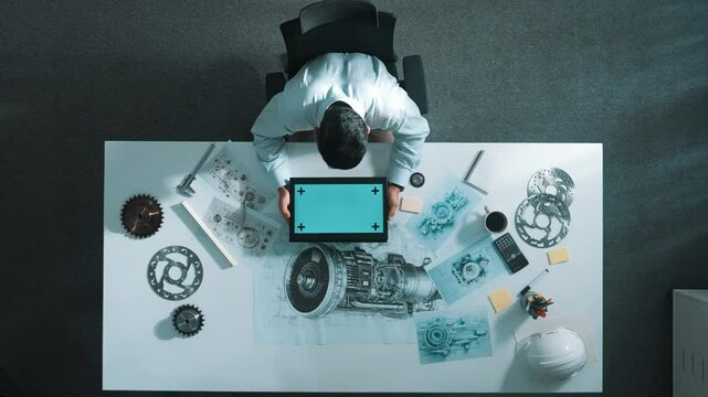 Top view of skilled engineer writing in paper while looking at tablet display green screen with turbine engine sketch design and metal gear and architectural equipment scatter around. Alimentation.