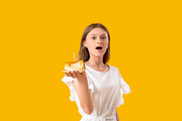 Surprised young woman with birthday cake on yellow background