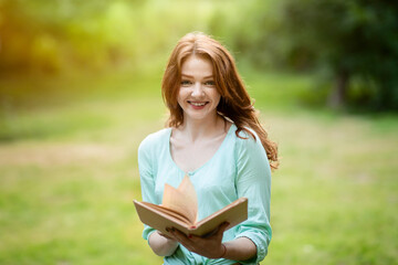 Fototapeta premium Beautiful Redhead Teen Girl Reading Book Outside In Summer Park, Posing At Camera And Smiling, Copy Space