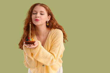 Beautiful young woman blowing out candle on birthday cupcake against green background
