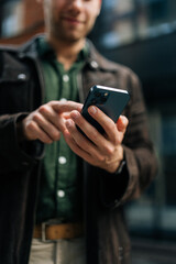 Vertical selective focus shot of unrecognizable man holding and using smartphone standing on city street, browsing internet, checking social media, using mobile app. Concept of modern communication.