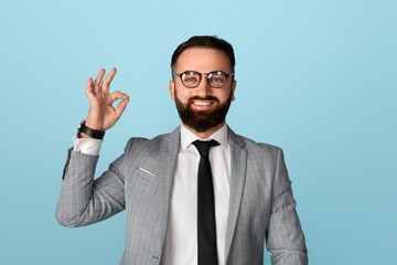 Happy businessman in formal suit showing okay gesture on blue studio background